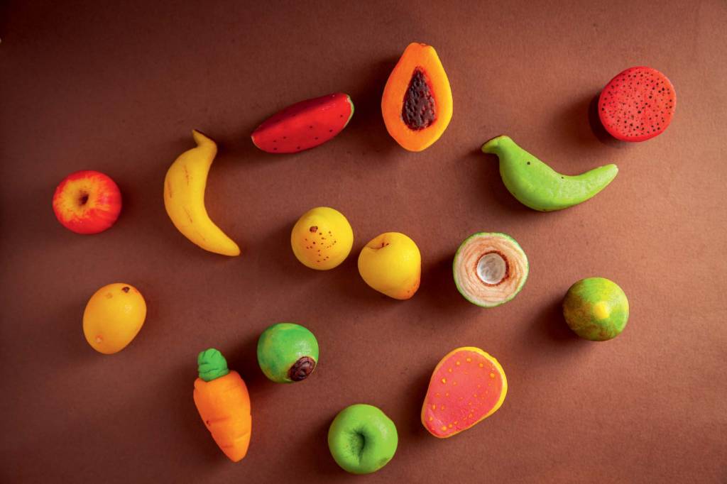 doces de marzipã no formato de frutas e legumes vistos de cima. Banana, mamão e cenoura são alguns deles