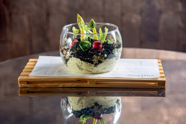 Recipiente de vidro em formato de terrário contendo legumes e espuma de aspargos verde que lembra a areia colorida dos terrários de verdade
