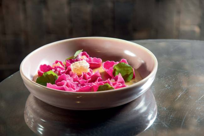 Prato de ceramica sobre superfície espelhada contendo massa recheada de tom rosado com bordas em formato que lembra pétalas de flor