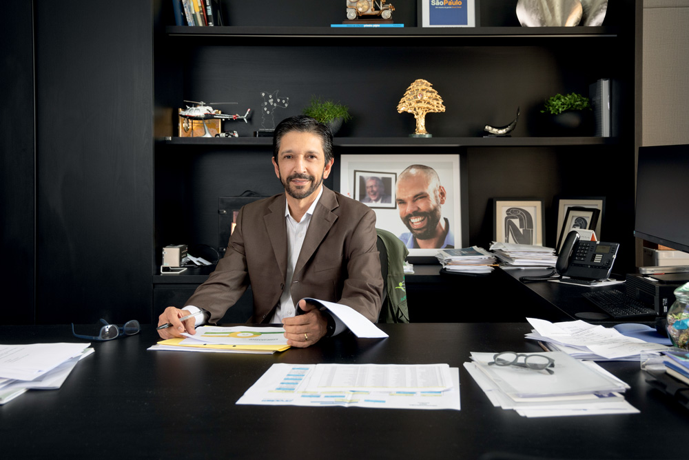 Homem vestindo blazer preto sentado em uma mesa preta e segurando papéis. Ele olha para a frente