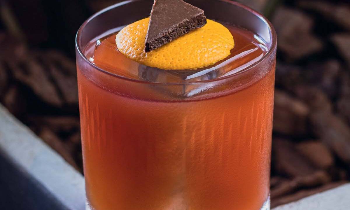 Copo baixo contendo drinque laranja avermelhado com cubo de gelo, rodela de casca de laranja e triângulo de chocolate em cima