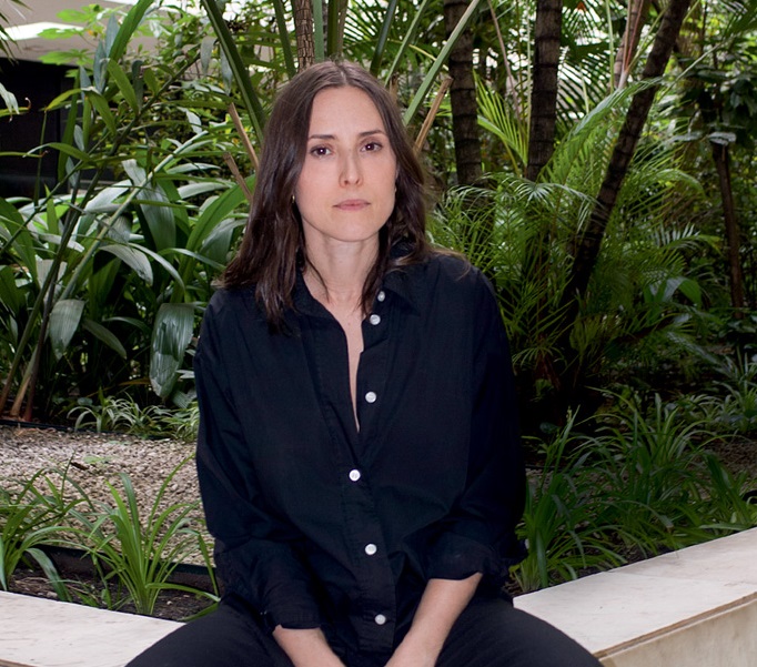 Foto de Verena Smit, usando camisa preta, com pose séria, em frente a banco com paisagem verde ao fundo