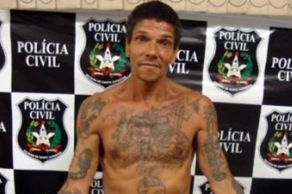 Foto de homem sem camisa, tatuado, diante de parede com logo da Polícia Civil