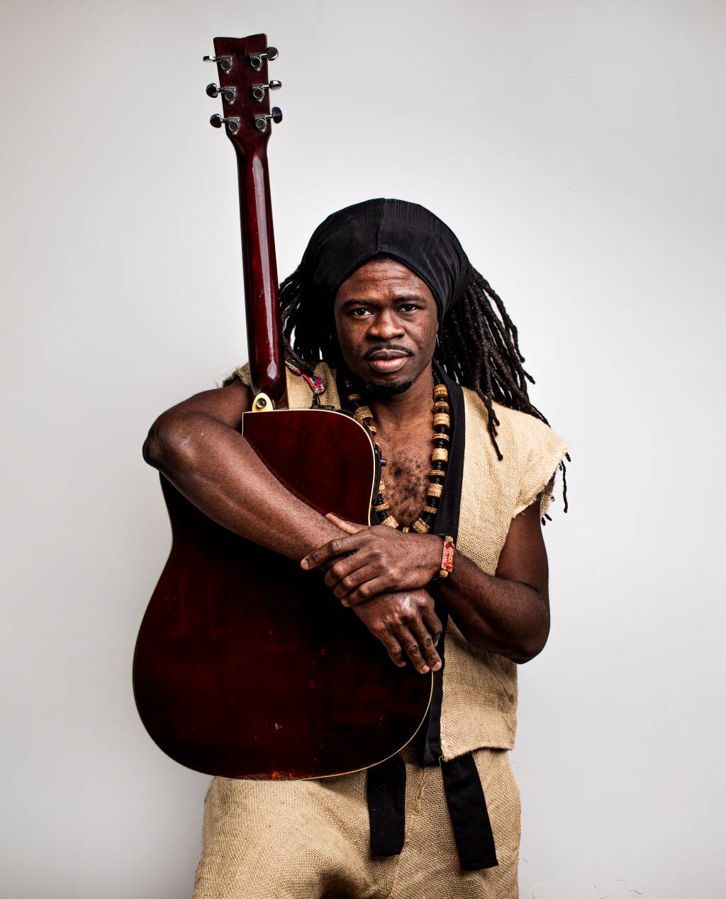 Cantor e compositor Yannick Delass posa abraçado com violão de pé. Tem dreads, pele negra e veste um colete cor de palha.