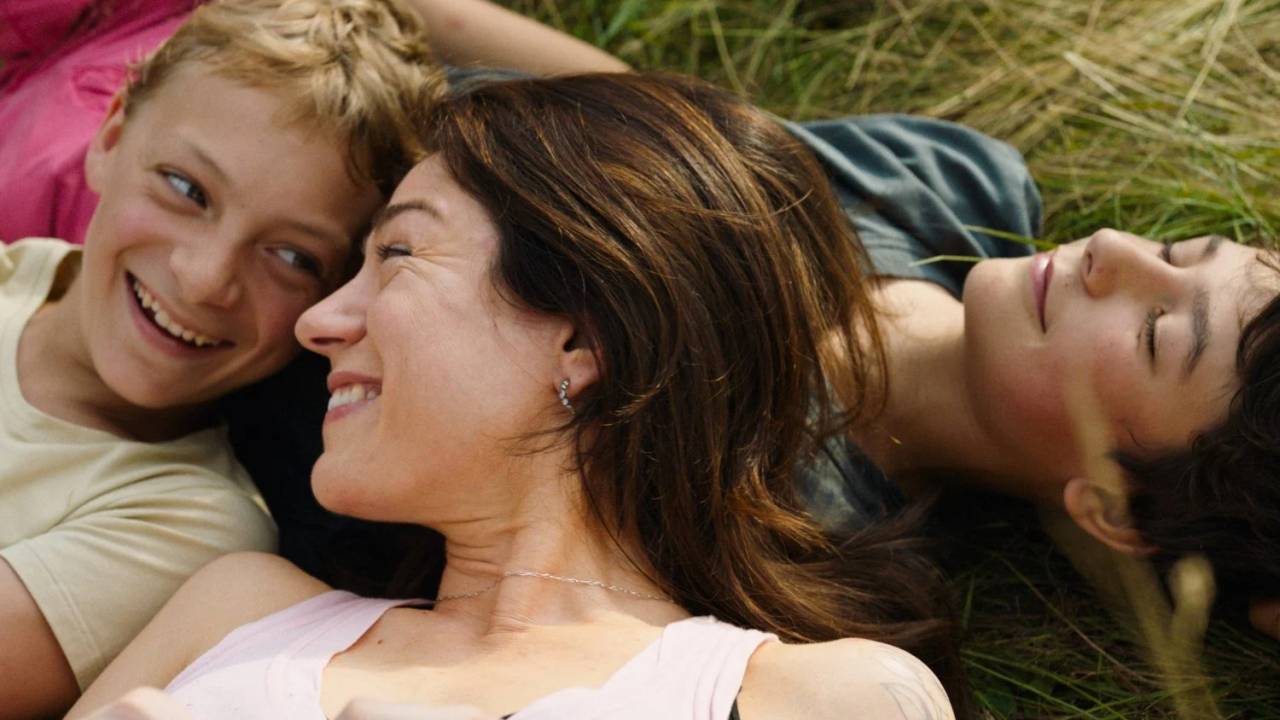 Mulher sorri no meio de dois meninos enquanto estão deitados na grama