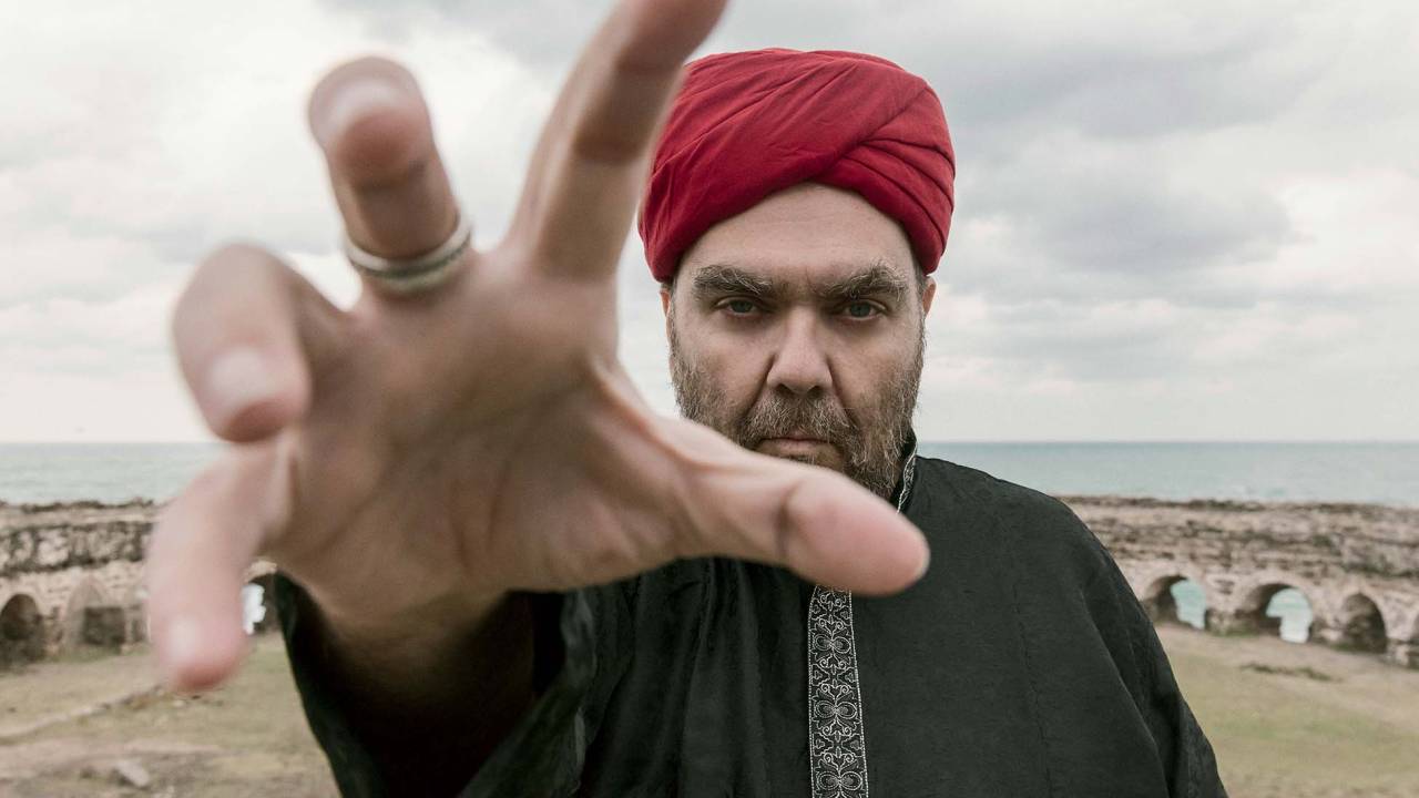Imagem mostra homem com turbante vermelho, estendendo e abrindo as mãos sobre a lente