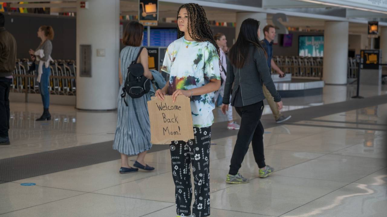 No aeroporto, menina está parada com uma placa de 'Bem-vinda, mãe'