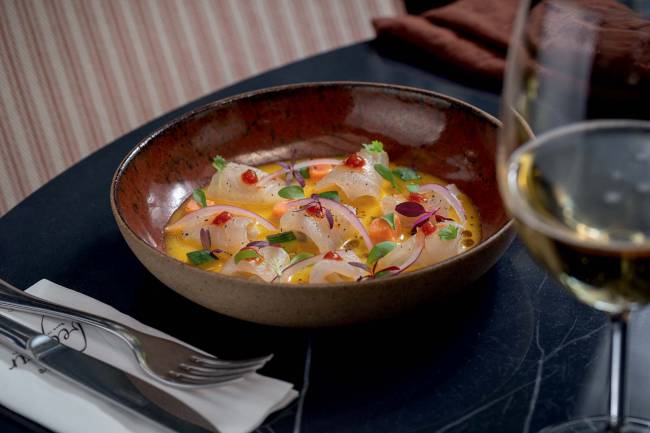 Cumbuca escura sobre mesa preta contendo ceviche de peixe branco, batata-doce, pepino, molho de maracujá e pimenta