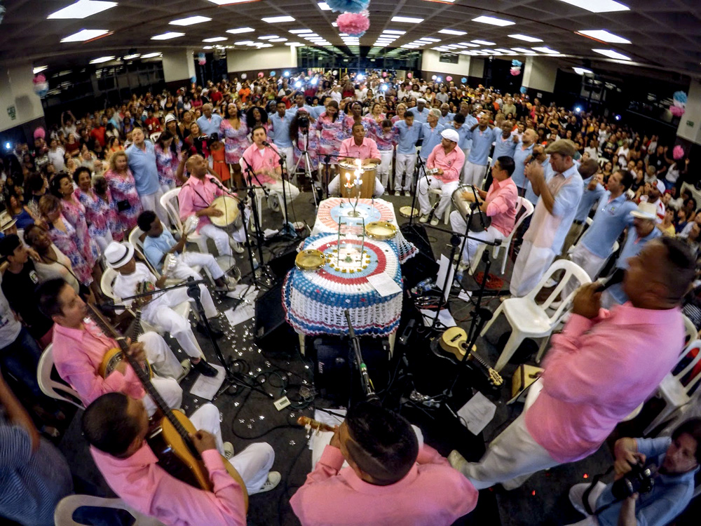 Imagem mostra roda de samba com sambistas vestindo camisa rosa