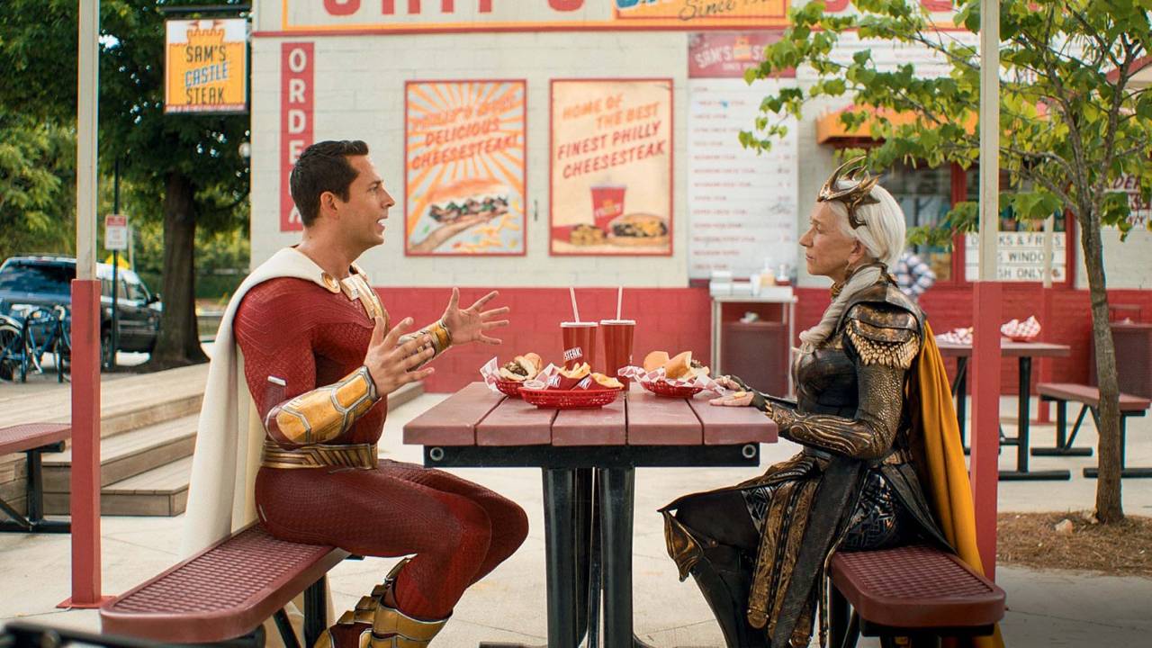 Imagem mostra super-herói com roupa vermelha sentado em frente a outra heroína. Eles estão comendo um fast food