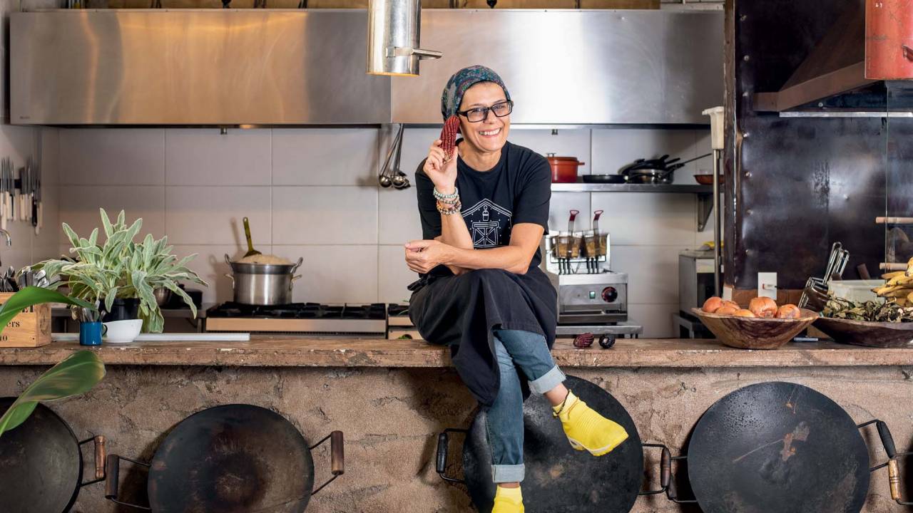 Angelita Gonzaga veste preto e bandana sentada no balcão da cozinha do restaurante