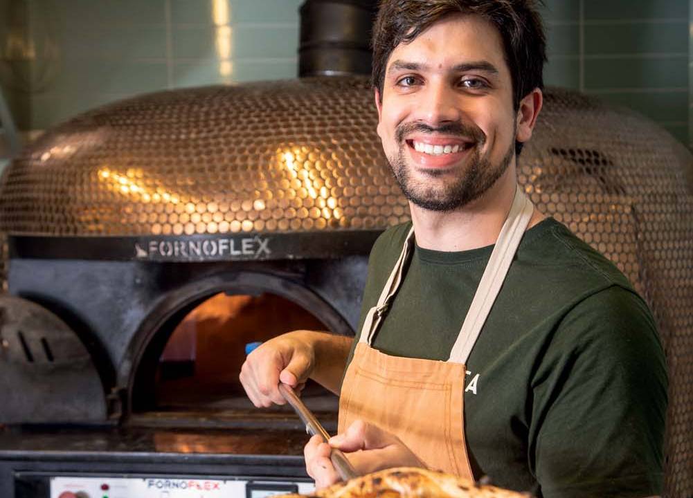 Homem de camiseta preta e avental, segurando pá para pizza com pizza, ao lado de forno de pizza