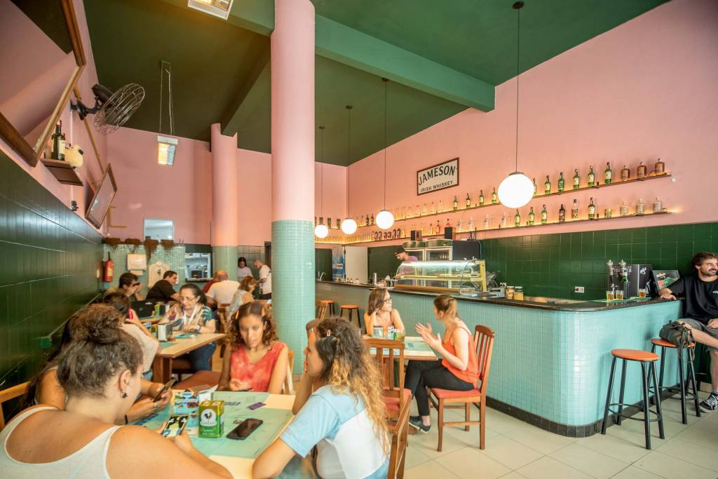 Ambiente interno do escarcéu com paredes cor-de-rosa, cobertas na metade inferior com azulejos verdes, com mesas de madeira e clientes sentados