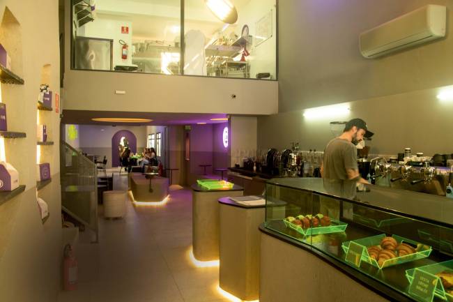 Ambiente interno do café com paredes cinzas, luzes roxas e bandejas verdes com pessoa trabalhando do lado direito