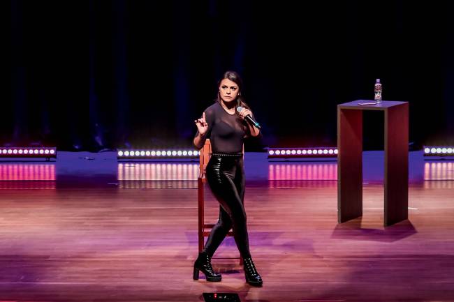 Bruna Louise Mulher branca de cabelos escuros fala em microfone sobre palco