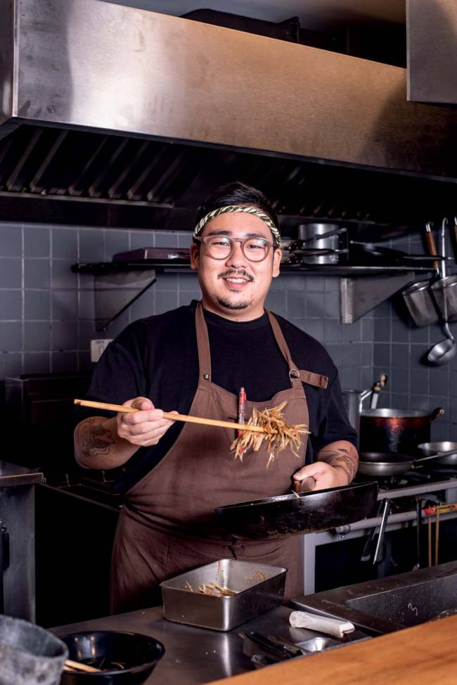 Chef asiático com blusa preta e avental marrom em cozinha segurando porção de comida em fatias com utensílio de cozinha