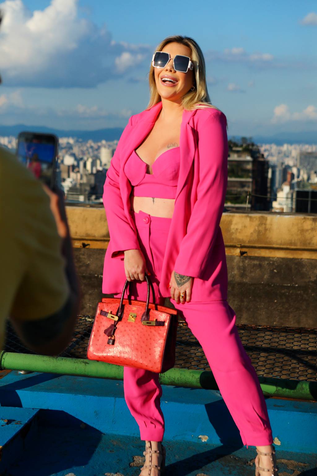 Mulher branca e loira posa no topo de prédio com roupas rosa pink e bolsa na mão direita.