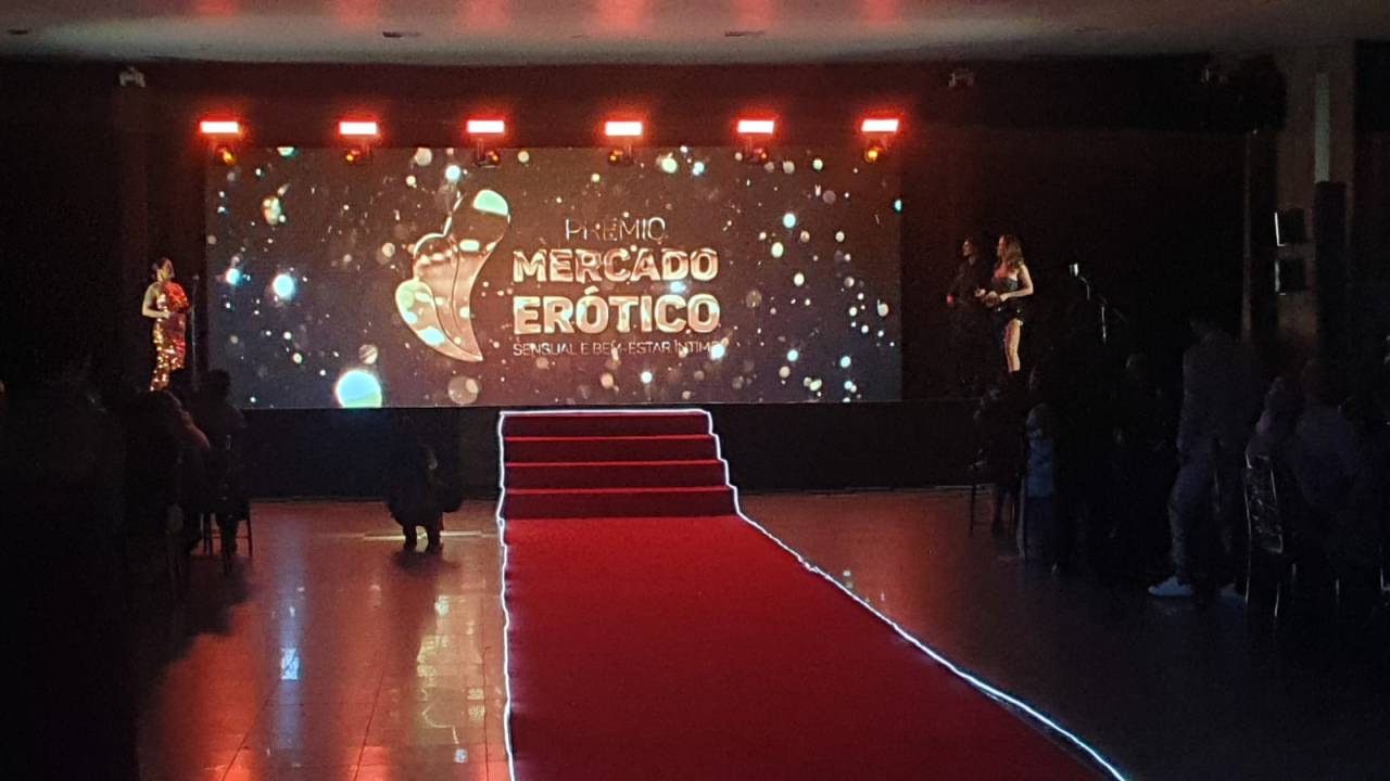 Tapete vermelho iluminado e palco com telão que diz "Prêmio Mercado Erótico".