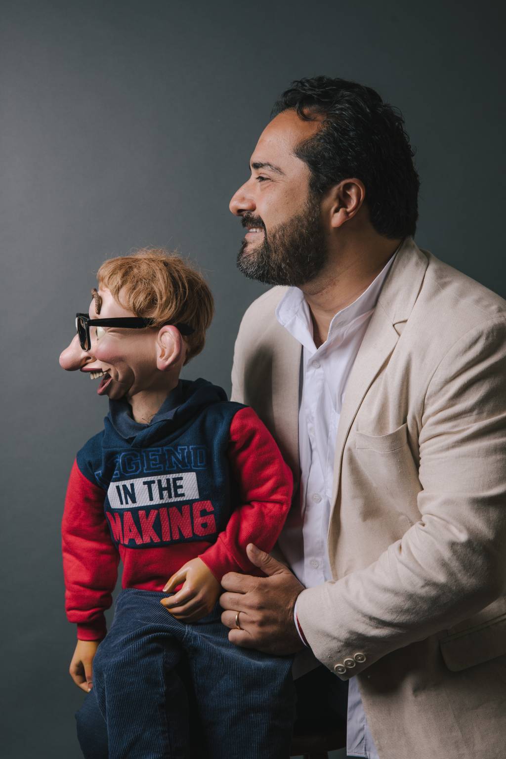 Homem branco de cabelos pretos posa olhando para o lado com boneco de ventriloquismo vestindo camiseta azul e vermelha com óculos de grau.