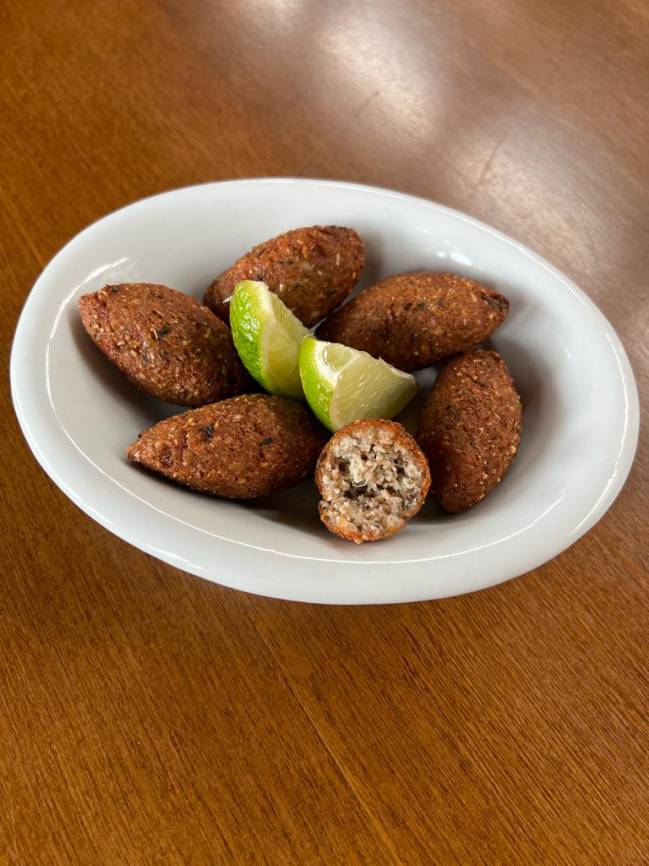 Porção de mini quibes de peixe em prato de porcelana, sobre mesa de madeira, com cunhas de limão