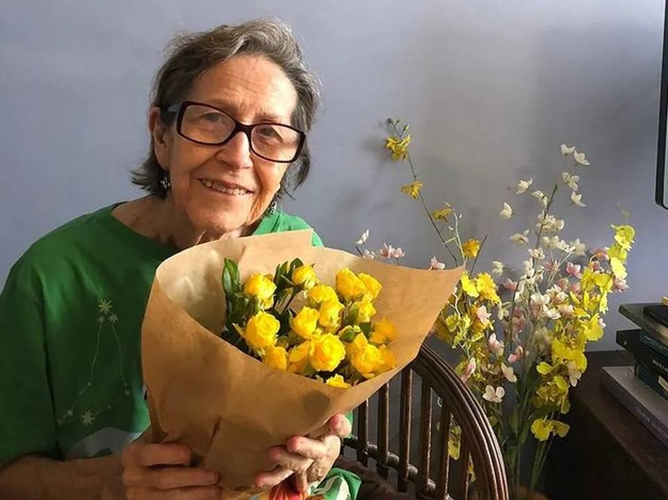 Foto de senhora com cabelo grisalho sentada sorrindo, com blusa verde e óculos, segurando buquê de flores amarelas