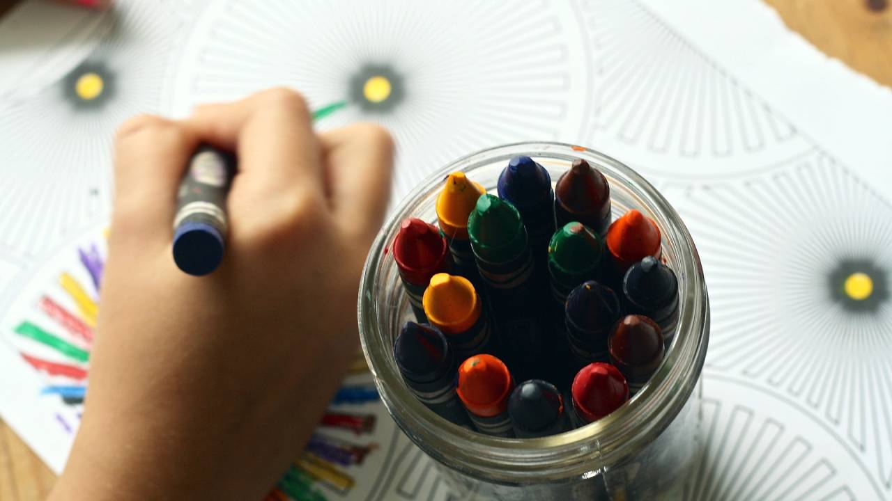 Foto mostra mão de criança desenhando ao lado de estojo com lápis de cores