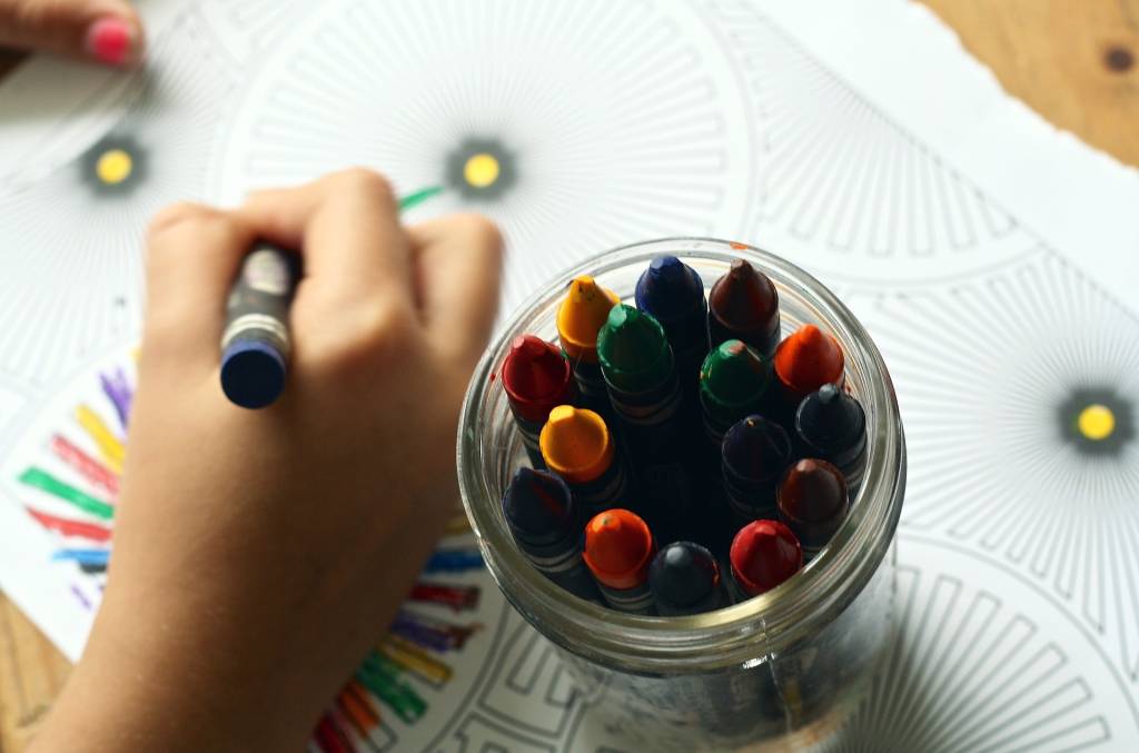 Foto mostra mão de criança desenhando ao lado de estojo com lápis de cores