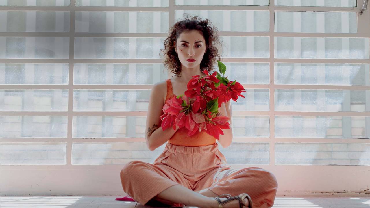 Com vestido de cores claras e flores nas mãos, mulher branca posa sentada de pernas cruzadas no chão.