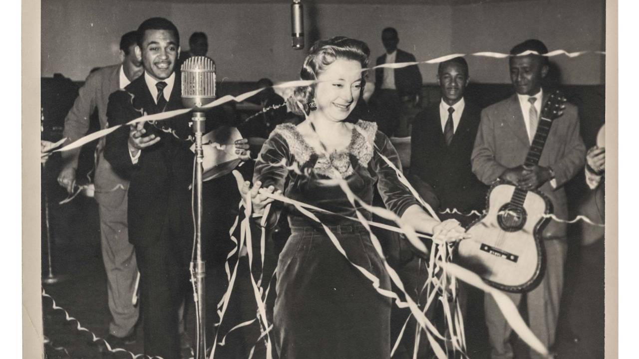 Imagem mostra foto em preto e branco com mulher em meio a círculo de homens com microfone em sua frente