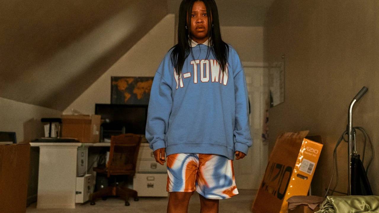 Menina de moletom azul parada, com feição séria, em um quarto claro