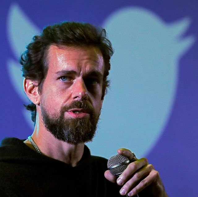 Um homem branco de barba e cabelos curtos segurando um microfone e o logo do Twitter, que é um pássaro azul, no fundo