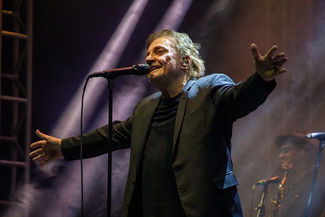 Um homem de terno e camiseta preta cantando ao microfone de braços abertos