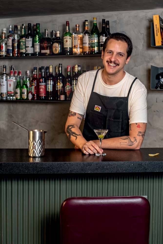 Homem branco sorrindo atrás de balcão de bar segurando um dry martini.