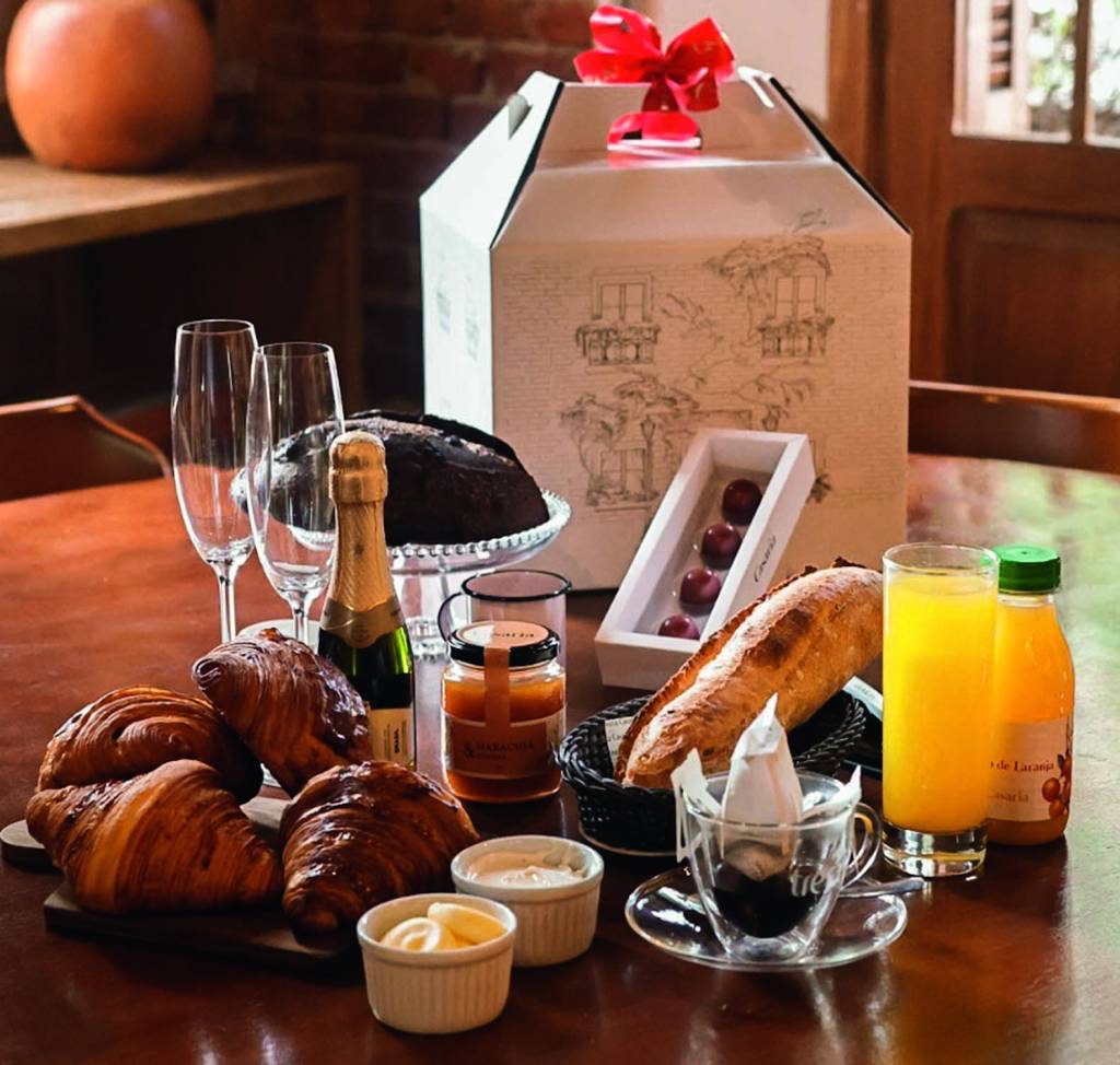 Mesa de madeira com croissants, taças e garrafa de espumante, pães, xícara de café, bombons e suco de laranja.