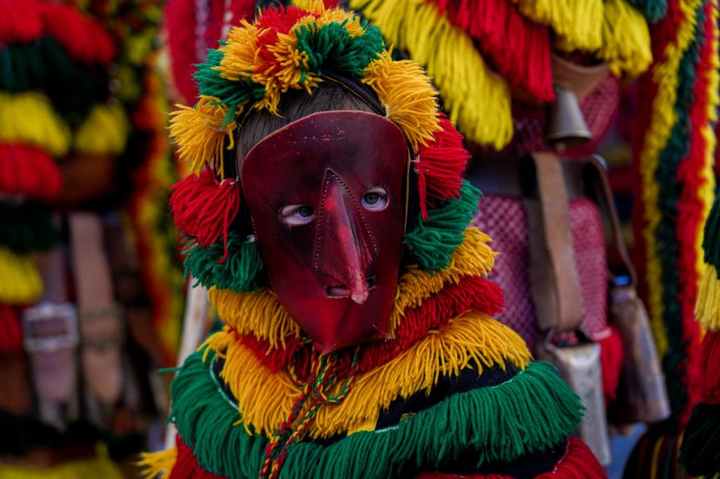 Torso de pessoa fantasiada com máscara de couro de nariz pontudo e roupa colorida com fios de lã em amarelo, vermelho e verde.