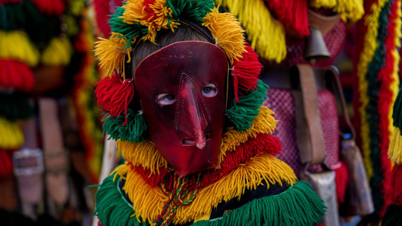 Torso de pessoa fantasiada com máscara de couro de nariz pontudo e roupa colorida com fios de lã em amarelo, vermelho e verde.