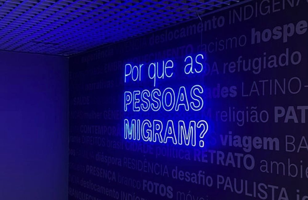 Letreiro de luz azul com a frase "Por que as pessoas migram?", na exposição do Museu da Imigração