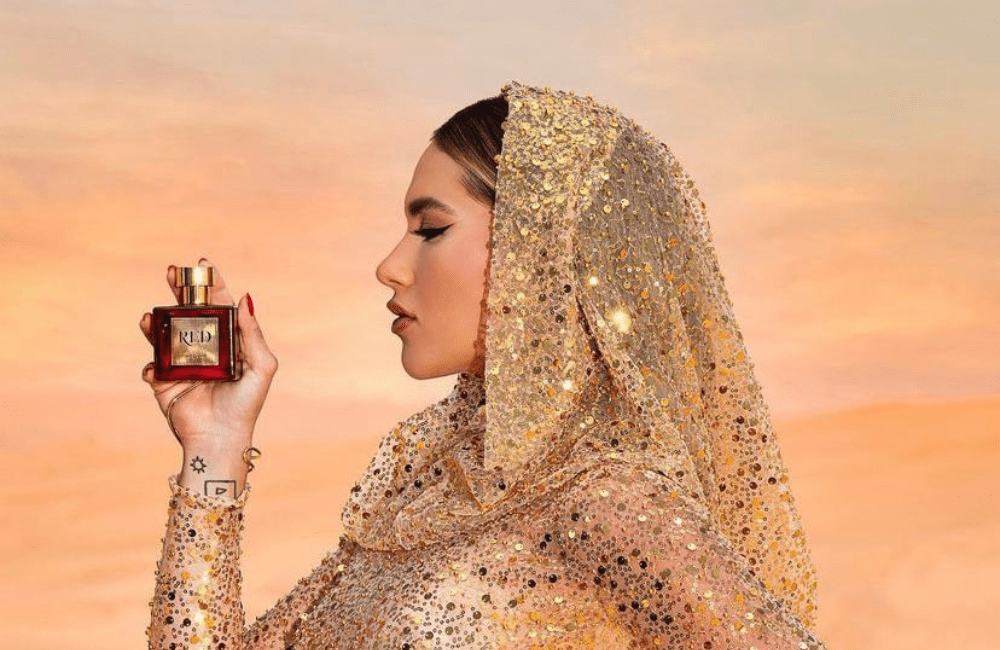 Virginia aparece de perfil, segurando um perfume de frasco vermelho, enquanto usa uma roupa brilhante, que remete as vestes de mulheres mulçumanas