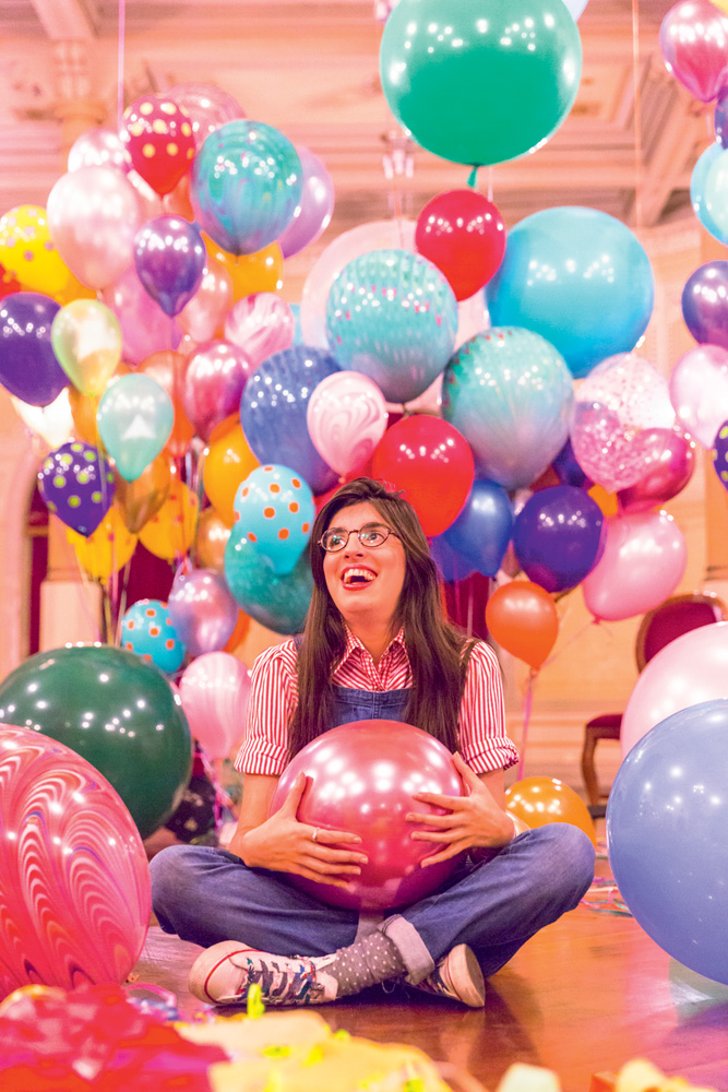Mulher branca de óculos e cabelos castanhos lisos aparece sentada de pernas cruzadas envolta por muitos balões coloridos de festa.