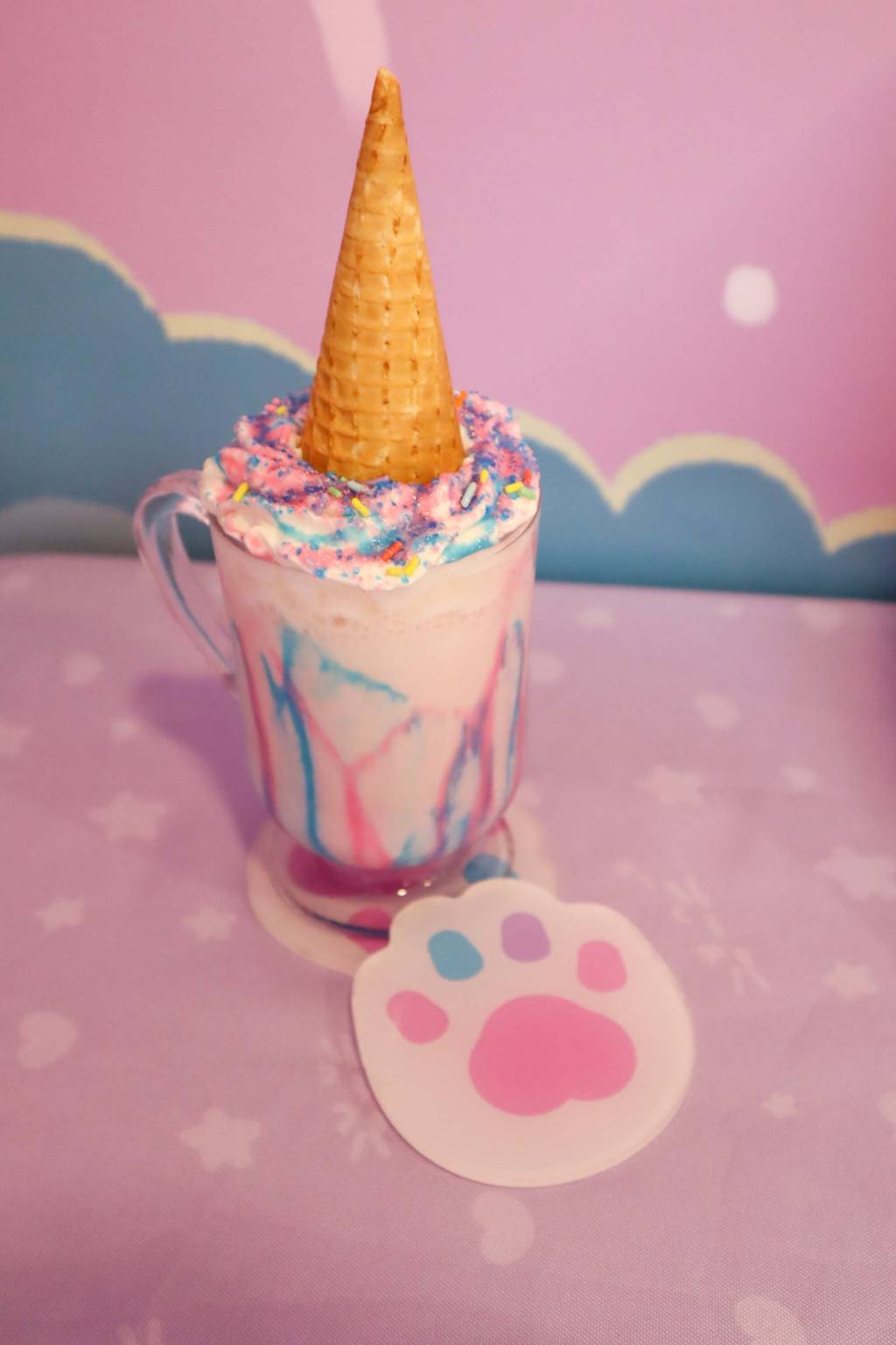 Caneca de vidro com bebida rosa, calda e granulados azul e rosa coberta por casquinha de sorvete.