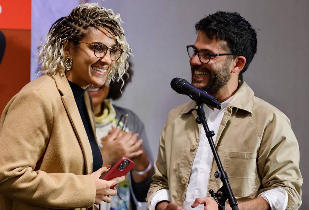 Mulher loira de óculos e homem de óculos com barba sorriem lado a lado, em frente a microfone preto.