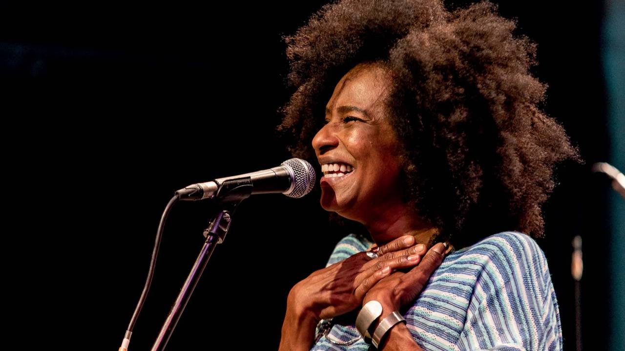 Mulher negra com black power canta em frente ao microfone com as mãos no peito.