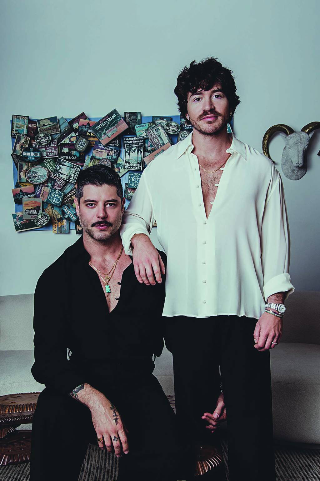 Dois homens brancos encaram a câmera, o da esquerda sentado com topete, bigode e camisa preta. O da direita de pé, apoiando o braço no ombro do outr, com cabelos escuros, barba e camisa branca com botões abertos no peito.