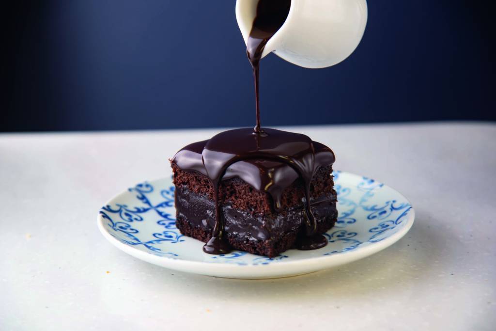 Prato de louça branca e azul com pedaço quadrado de bolo de chocolate com calda sendo derrubada por cima em mini jarra.