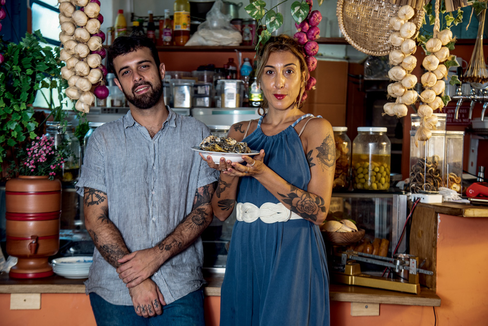 Homem e mulher brancos de braços tatuados a frente do balcão alaranjado do bar Manu.el.