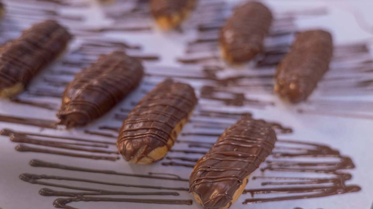 Mini eclairs alinhadas em diagonal sobre tampo branco "manchado" de linhas de chocolate.