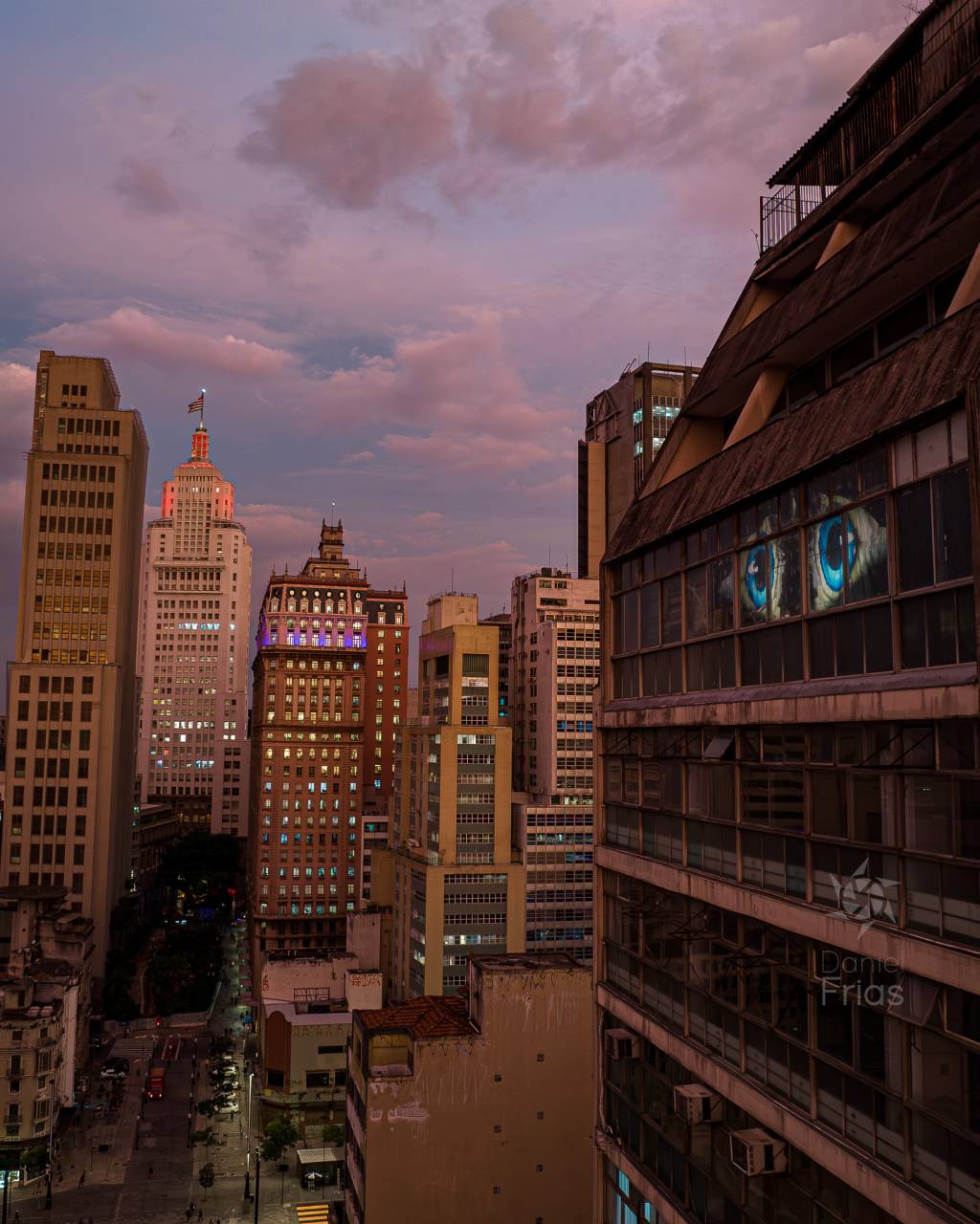 Foto exibe prédios com horizonte em fim do dia. Um deles tem duas janelas decoradas com olhos gigantes iluminados.