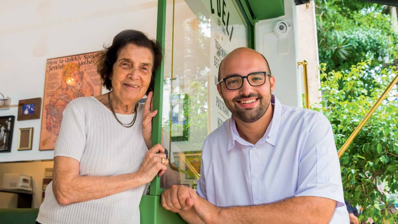 Senhora e homem brancos de camisetas brancas apoiados na fachada do restaurante Z Deli.