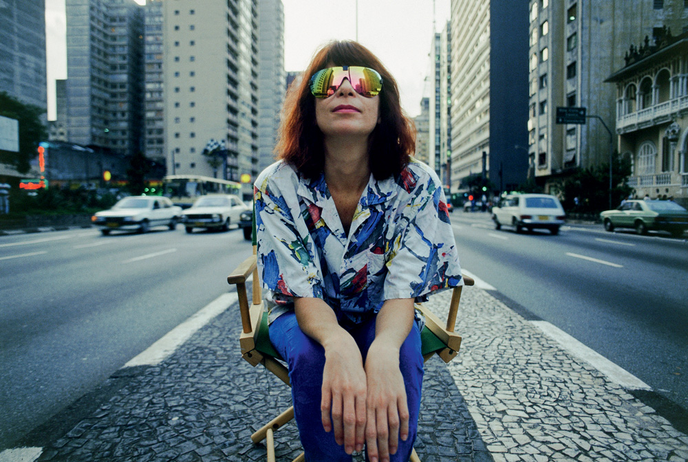 Imagem mostra mulher de cabelos vermelhos sentada, com óculos escuros, em calçada entre avenidas