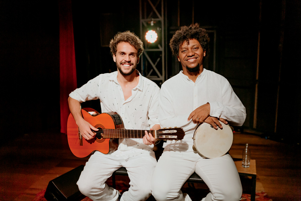 Dois homens sorriem sentados lado a lado, um com um violão no colo e outro com um pandeiro. Ambos vestem roupas brancas.
